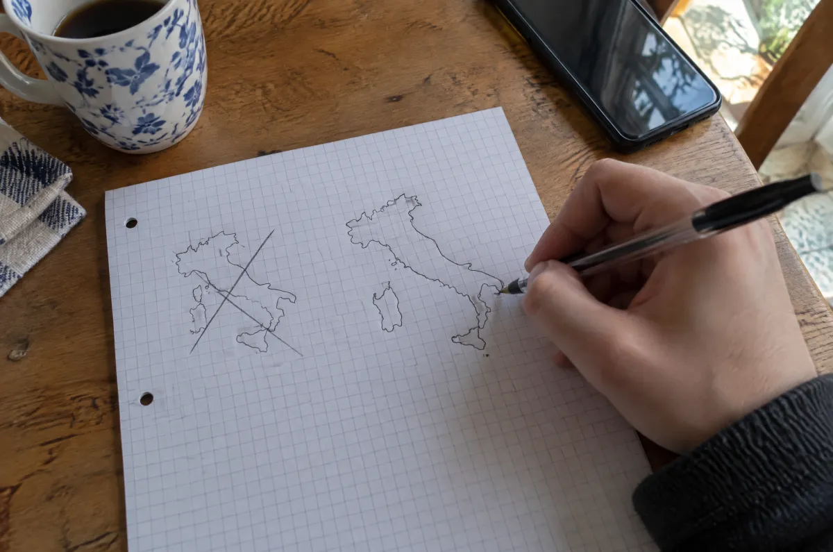 A hand drawing the outline of Italy from memory on squared notebook paper next to a crossed-out first try.